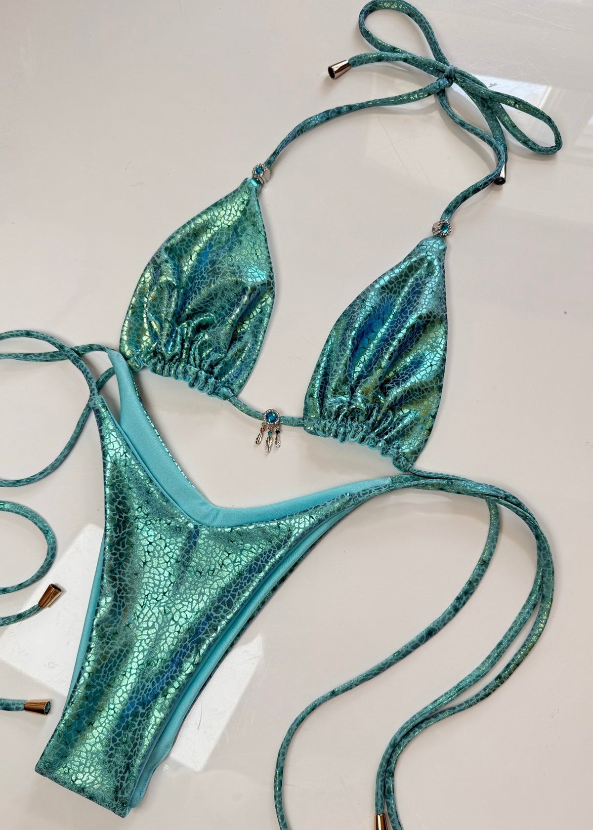 Shiny turquoise bikini set with textured pattern and beaded charm, laid flat on white surface