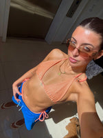 Woman in peach bikini top and blue swim shorts wearing sunglasses, standing indoors in sunlight