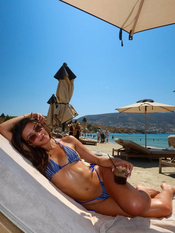 Woman in blue bikini lounging on beach chair under umbrella, holding drink, seaside view