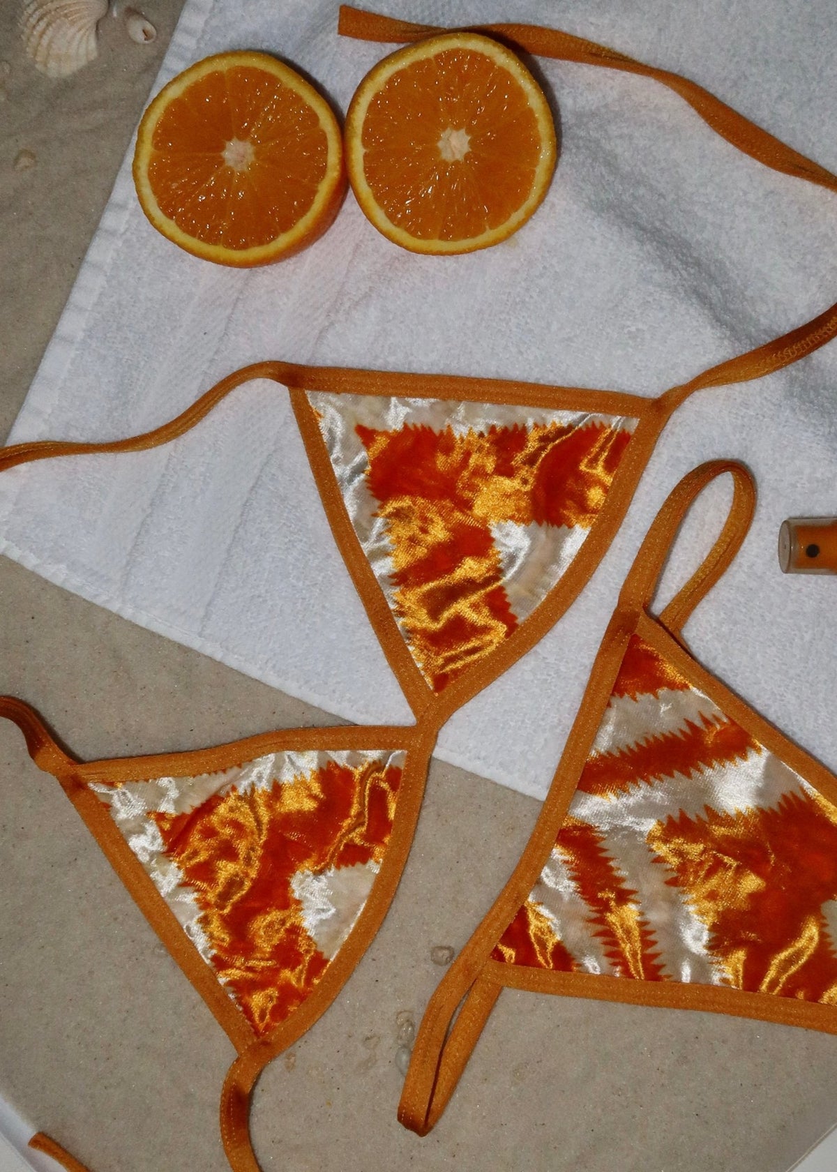Handmade orange and white velvet bikini set on towel, with fresh orange halves on sand