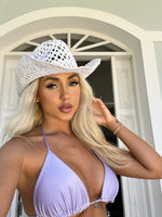 Woman in lavender bikini top and white cowboy hat posing outdoors in front of a white archway