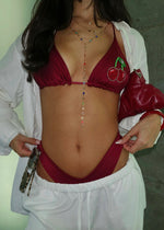 Woman wearing a burgundy bikini with beaded cherry detail, layered gold necklaces, and white cover-up.
