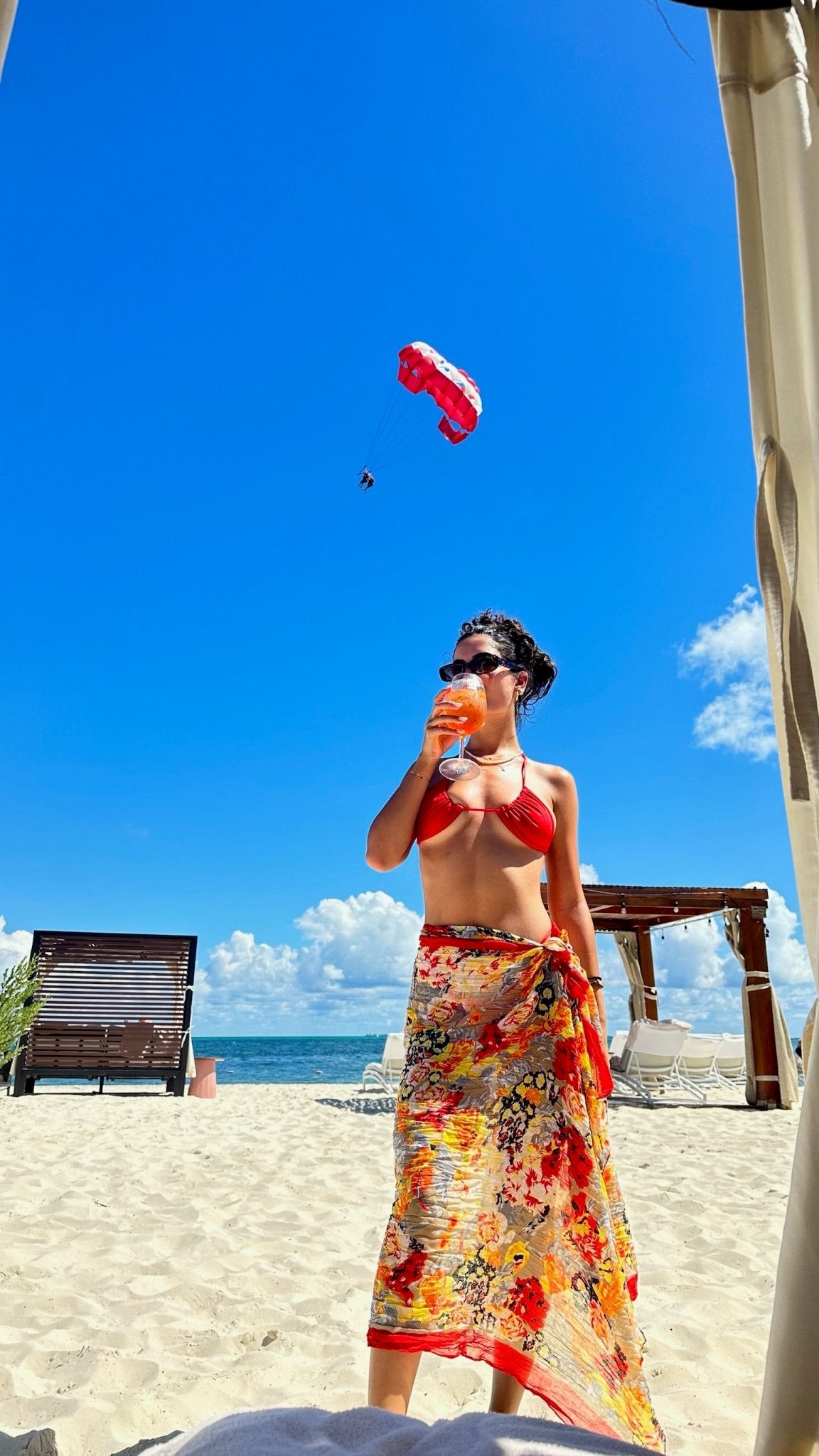 Woman in red bikini and floral sarong enjoying a drink on a sunny beach, clear blue sky
