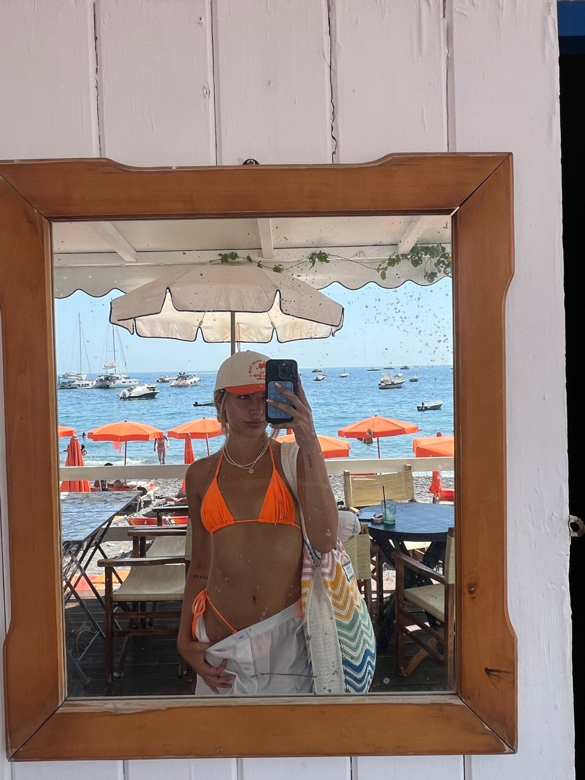 Woman in orange bikini with beachwear and tote bag, reflected in mirror at seaside cafe with umbrellas and boats.