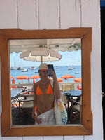 Woman in orange bikini with beachwear and tote bag, reflected in mirror at seaside cafe with umbrellas and boats.