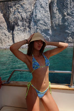 Woman in patterned blue and green bikini posing on a boat by clear water and rocky cliffs