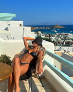 Woman in black bikini and sun hat relaxing on balcony with ocean view, summer swimwear style