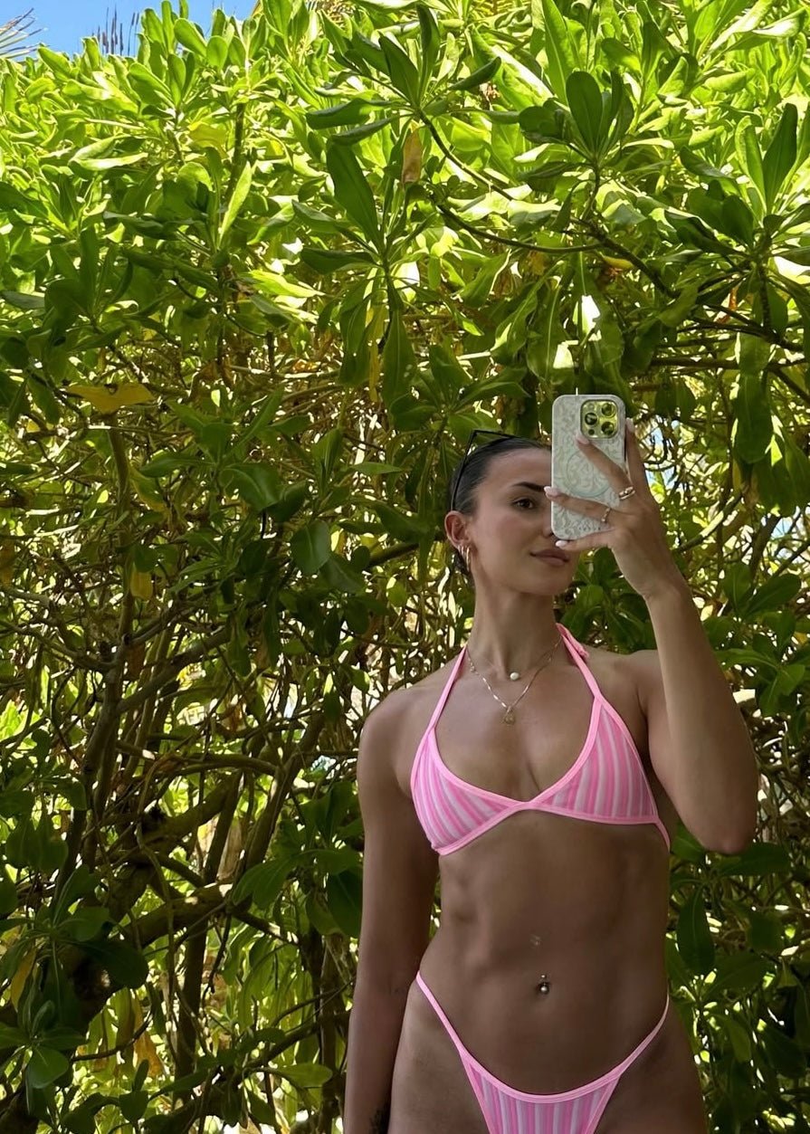 Woman in pink and white striped bikini taking selfie outdoors in front of green leaves