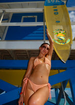 Woman in handmade peach bikini posing by blue and yellow beach lifeguard hut in Florida