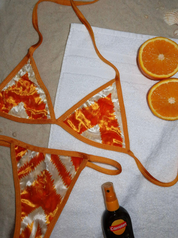 Orange and cream handmade bikini set on sand with a towel, tanning oil, and orange slices