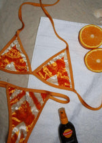 Orange and cream handmade bikini set on sand with a towel, tanning oil, and orange slices