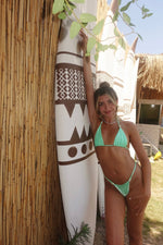 Woman in a handmade mint green bikini posing by surfboards and bamboo hut outdoors