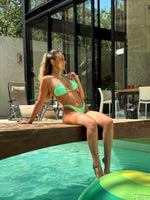 Woman in green bikini sitting poolside outdoors, modern glass house in background