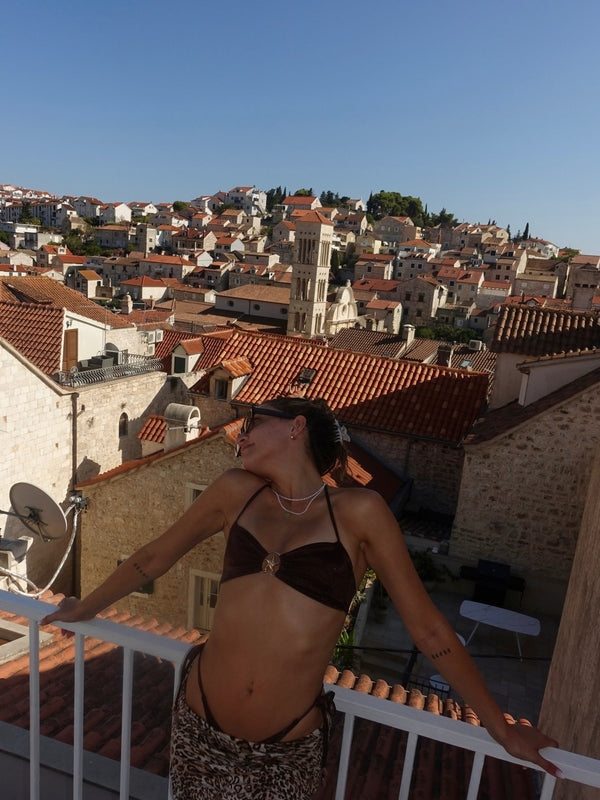 Woman in handmade bikini top and animal print skirt posing on balcony with Mediterranean town view