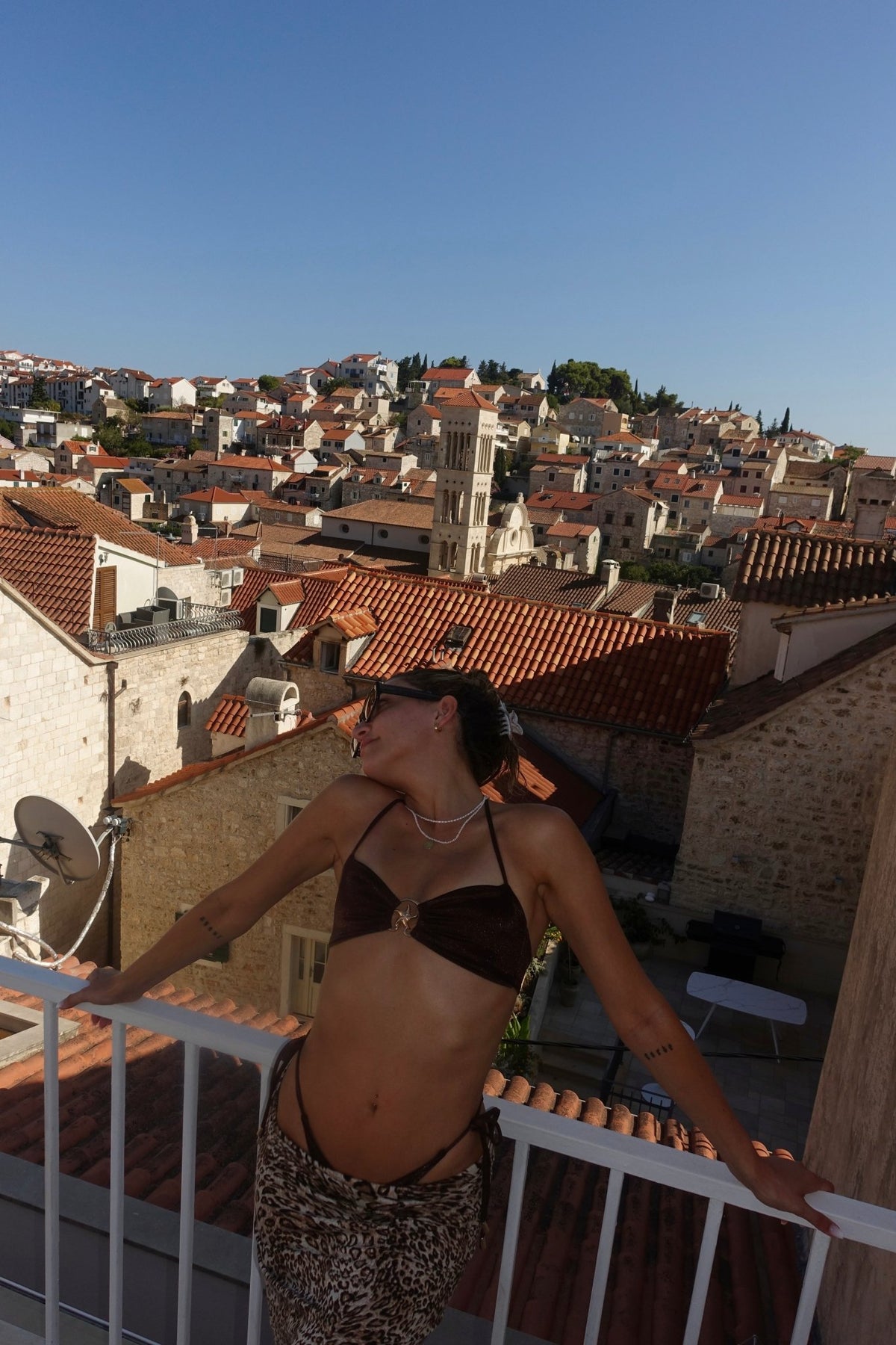 Woman in handmade bikini top and animal print skirt posing on balcony with Mediterranean town view
