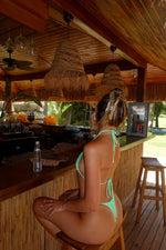 Woman in mint green bikini sitting at tropical bar with wooden decor and woven pendant lights