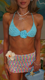 Woman wearing a blue crochet bikini top with flower, sequin mini skirt, and holding blue bag