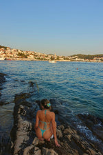 Woman in handmade turquoise bikini sitting on rocky shore by the sea with ocean view