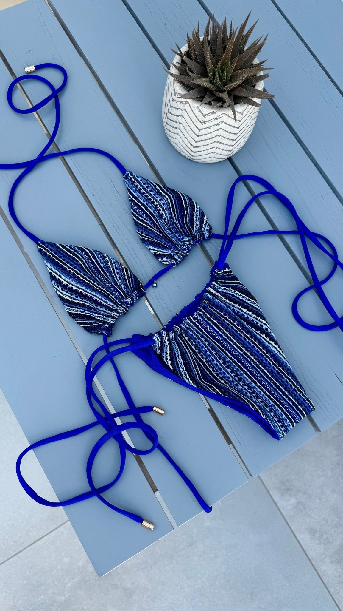 Blue striped handmade bikini set with tie straps on a blue table beside a potted succulent