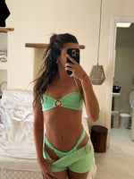Woman wearing mint green bikini with shell detail and crochet shorts, taking mirror selfie indoors