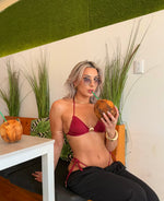 Woman in red bikini top and black pants drinking coconut, seated in tropical themed cafe.