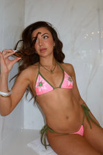 Woman wearing pink and green floral bikini set, holding sunglasses, sitting in a bright tiled room.