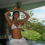 Woman in a white and mint green bikini top by a pool, holding a pineapple crown on her head, surrounded by lush greenery.