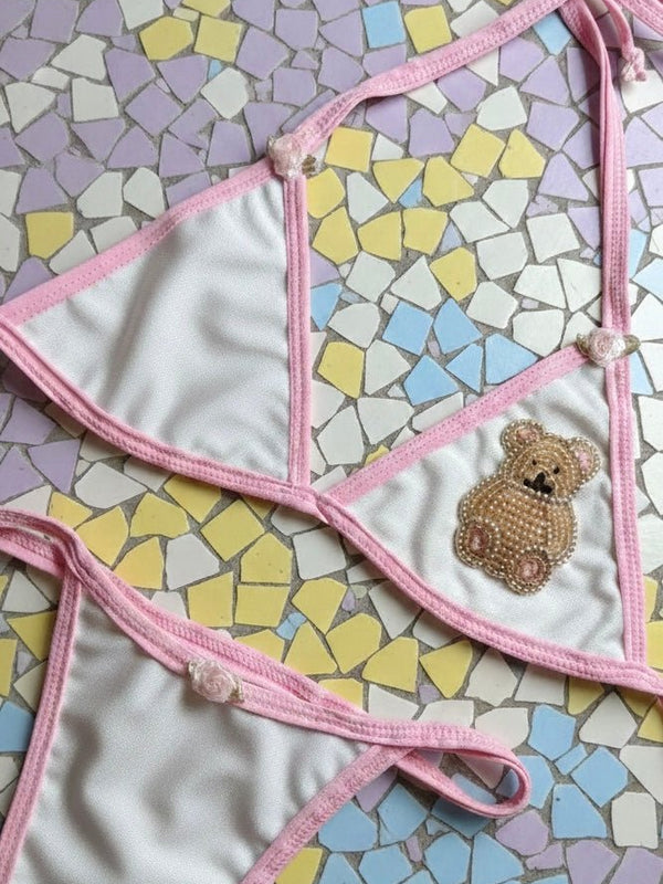Handmade white and pink bikini set with beaded teddy bear patch, laid on pastel mosaic tiles
