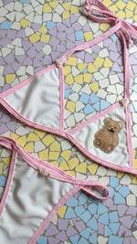Handmade white and pink bikini set with beaded teddy bear patch, laid on pastel mosaic tiles