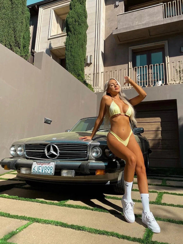 Woman in a yellow bikini posing by a vintage Mercedes car in a sunny backyard