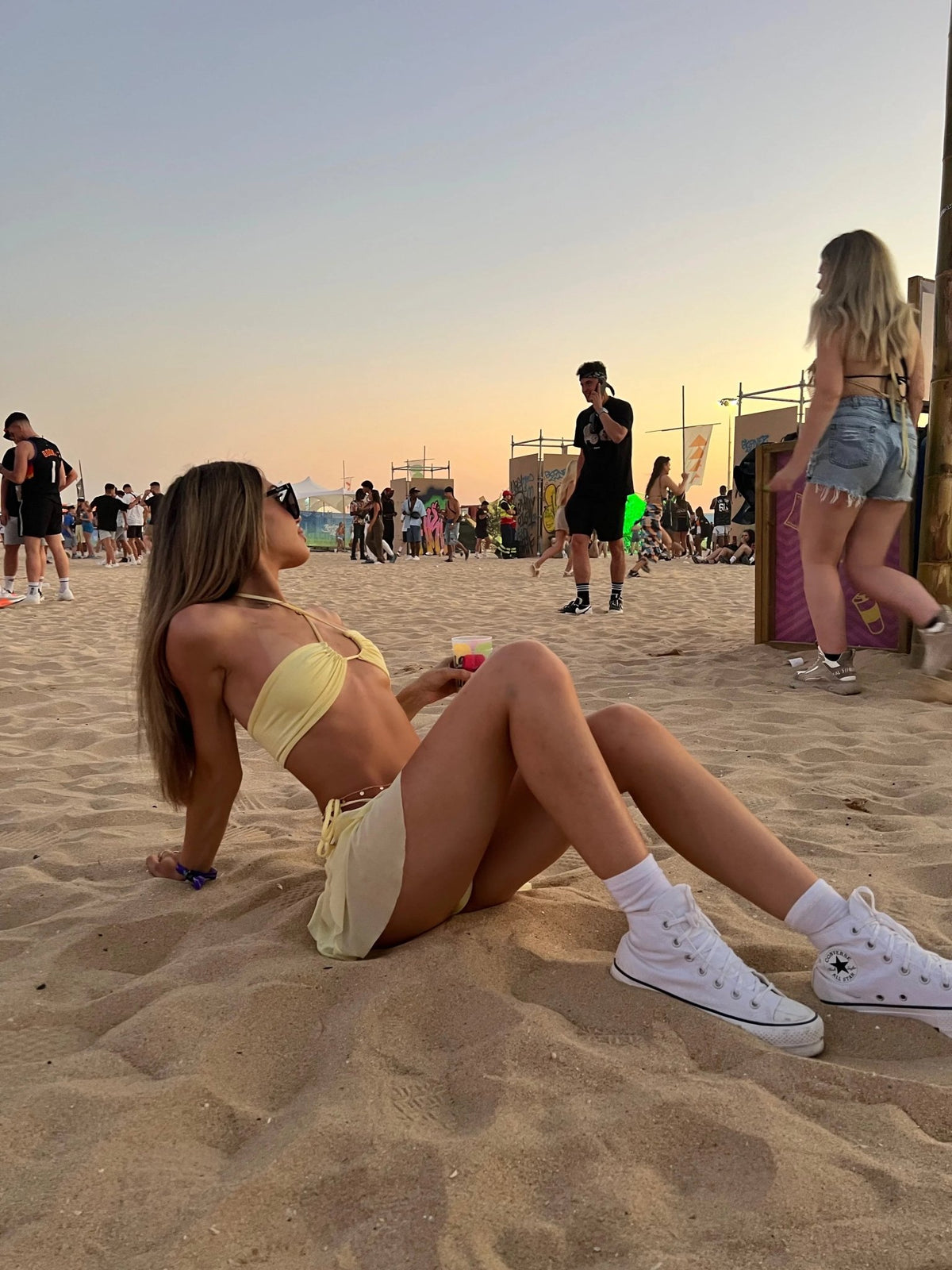 Woman in yellow bikini set lounging on sandy beach at sunset, surrounded by festival crowd